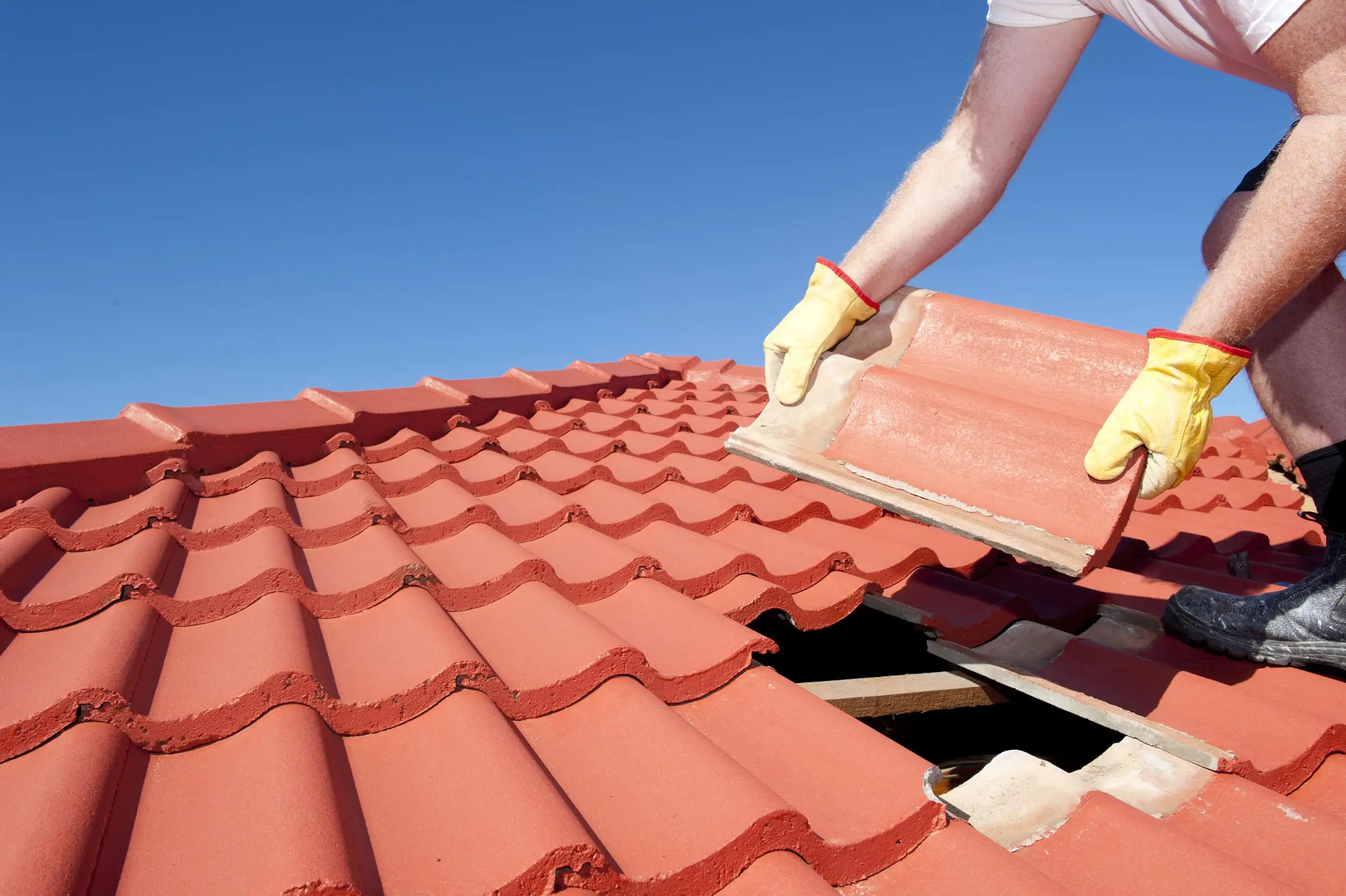 Roof Repair, Worker With Yellow Gloves Replacing Red Tiles Or Shingles On House With Blue Sky As Background And Copy Space.